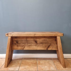 May include: A rustic wooden bench featuring a rectangular seat and angled legs. The bench is constructed from natural brown timber with visible wood grain, knots, and a smooth finish, showcasing a sturdy design with a horizontal support panel.