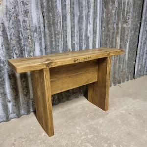 May include: A small, rustic wooden bench with a natural finish. The bench has a rectangular seat and four sturdy legs. The text "RSI 2020" is stamped on the top. The bench is suitable for indoor or outdoor use.