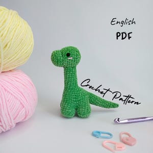 Może przedstawiać: Zielona pluszowa zabawka amigurumi dinozaura z czarnymi oczami. Wzór na szydełku PDF w języku angielskim.