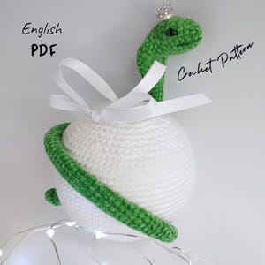 May include: A green crocheted snake with a crown on its head wraps around a white crocheted sphere. The snake is holding a white ribbon with a bow. The text "English PDF" and "Crochet Pattern" is visible in the image.