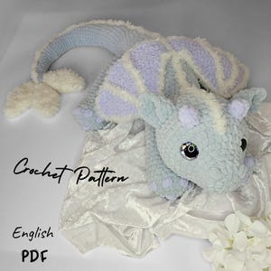 May include: Crochet pattern for a light blue and purple dragon amigurumi toy. The dragon has a long tail and large eyes. The pattern is available in English PDF format.