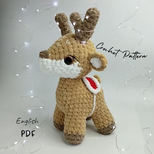 May include: Crocheted reindeer amigurumi toy with brown and white fur, red and white accents, and a brown nose. The toy is standing on a white surface with white string lights in the background. The text "Crochet Pattern" is visible in the upper right corner of the image. The text "English PDF" is visible in the lower left corner of the image.