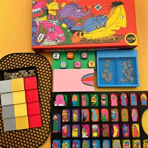 Peut inclure: Un jeu de société intitulé Athlètes de Compète comprenant une boîte rouge illustrée de personnages, un plateau noir avec des jetons colorés, un plateau bleu avec des étoiles, un porte-cartes rose et des dés numérotés de diverses couleurs.