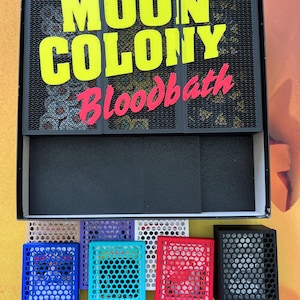 Könnte beinhalten: Eine Brettspielbox mit dem Titel "MOON COLONY Bloodbath" in gelben und rosa Buchstaben. Die Box enthält mehrere farbige Spielsteine in Blau, Lila, Weiß, Rot und Schwarz.