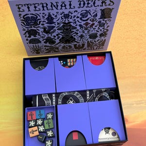 Peut inclure: Boîte de jeu de société ouverte, intitulée "ETERNAL DECKS" en noir sur un fond lavande. Contient des cartes, des dés et un tissu à motifs noir et blanc. La boîte est posée sur une surface jaune et orange.