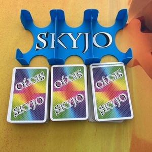 Può includere: Un porta carte in plastica blu con la scritta "SKYJO" in bianco. Tre mazzi di carte con un disegno arcobaleno e la scritta "SKYJO" sono davanti al porta carte. Lo sfondo è di colore arancione chiaro.