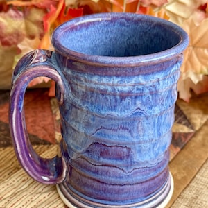May include: A ceramic mug featuring a cylindrical body with horizontal ridges and a curved handle. The exterior is finished with a glossy glaze in shades of deep purple and vibrant blue, creating a marbled effect that transitions to a white base.
