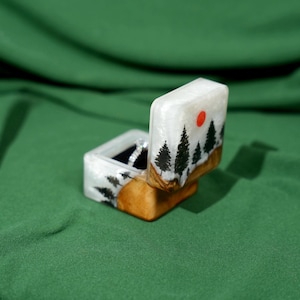 May include: A handcrafted ring box with a landscape design. The box is white with a resin finish, featuring black trees, a red sun, and a wooden base. The box is open, revealing a ring inside.