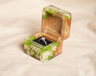 May include: Open wooden ring box with a clear resin exterior. The resin is decorated with white flowers and green foliage. Inside, a silver ring with a diamond sits on a black velvet cushion. The box is open, revealing the ring.