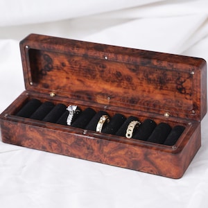 May include: A wooden jewelry box with a burl wood finish, open to reveal black velvet ring holders. Three rings are visible: a diamond engagement ring and two gold bands. The box is set against a white background.