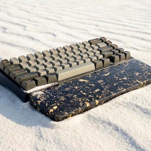 Peut inclure: Un clavier mécanique avec des touches grises et une base noire, posé sur une surface sablonneuse. Le repose-poignet est noir avec des éclats dorés. Le clavier a un design minimaliste, axé sur la fonctionnalité et l'esthétique.