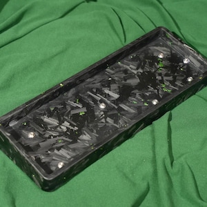 May include: A rectangular black tray with a carbon fiber pattern and several silver-colored rivets. The tray has a dark green interior and is set against a green fabric background. The tray is likely for organizing small items.