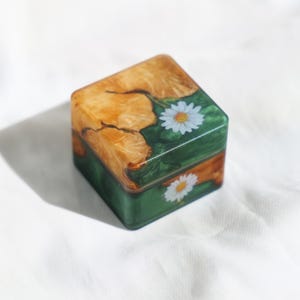 May include: Square wooden box with a natural wood grain top and green sides. The box features a white daisy flower design on the green sides. The top has a light brown and yellow wood grain pattern.