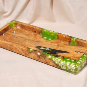 May include: A rectangular wooden tray featuring a natural wood grain base with integrated bright green resin sections. The resin contains embedded small white flowers and moss-like textures. Three small silver metal screw heads are visible.
