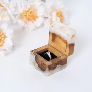 Könnte beinhalten: Geöffnete, quadratische Ringbox aus Holz und Kunstharz mit einem silbernen Ring. Die Box hat Goldblatt-Akzente. Der Ring hat einen großen, klaren Mittelstein. Auf weißem Untergrund, mit weißen Blumen im Hintergrund.