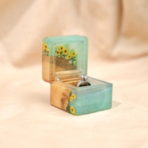 May include: A square ring box made of light-toned wood and translucent teal resin. The box features an embedded sunflower design on the exterior and interior lid. A silver-toned ring with a clear stone is positioned inside the open box.