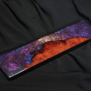 May include: A long, rectangular wooden board with a galaxy-themed epoxy resin inlay. The resin is a deep purple with silver glitter and a blue swirl. The wood is a reddish-brown color.