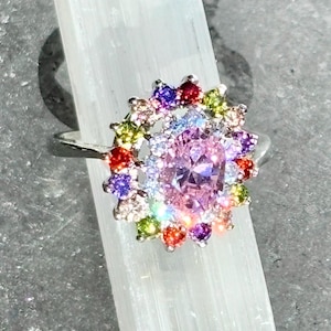 May include: A silver ring featuring a large, oval-cut pink gemstone surrounded by a halo of smaller, colorful gemstones. The ring's design is set against a white background.