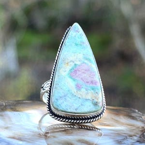 May include: A silver ring featuring a large, triangular gemstone. The stone has a light blue base color with pink and cream-colored inclusions. The ring's setting has a twisted rope design around the stone and decorative details on the band.