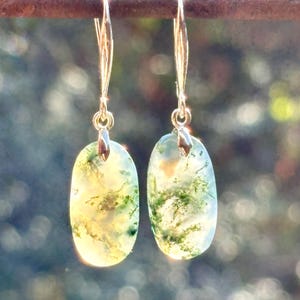 May include: A pair of drop earrings featuring oval-shaped moss agate stones with translucent white bases and intricate green dendritic inclusions. The stones are suspended from thin, polished silver-toned metal ear wires with a simple hook design.