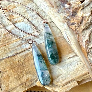 May include: A pair of drop earrings featuring teardrop-shaped moss agate stones with green inclusions suspended from curved copper-toned wire hooks. The stones are translucent with visible moss-like patterns throughout the polished surfaces.