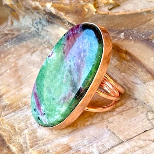 May include: A ring featuring a large, oval-shaped polished gemstone with a mottled green and deep magenta surface. The stone is set in a smooth, polished copper bezel with a triple-wire band design that wraps around the finger in a layered style.