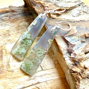 May include: A pair of rectangular earrings with a silver hook. The earrings are made of a translucent material with a mix of gray, white, and green patterns. The earrings are displayed on a wooden surface.
