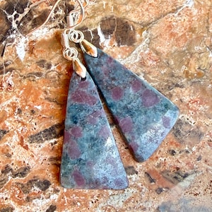 May include: A pair of gemstone earrings with a triangular shape. The earrings feature a silver hook and a stone with a blue-green base color and red spots. The earrings are displayed on a brown and orange marble surface.