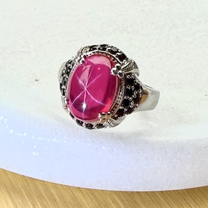 Könnte beinhalten: Ein silberner Ring mit einem großen, ovalen, rosa Edelstein mit sternförmigem Muster. Der Edelstein ist von kleineren schwarzen Steinen und aufwendigen silbernen Details umgeben. Ein einzigartiges Schmuckstück.
