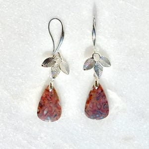 May include: A pair of silver-toned dangle earrings featuring a teardrop-shaped stone with a mottled reddish-brown and white pattern. Each earring is suspended from a curved wire hook and accented with three small, textured silver-toned leaf charms.