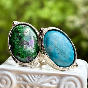 Puede incluir: Dos anillos de plata con piedras preciosas ovaladas. Uno presenta una piedra verde con inclusiones moradas, el otro una piedra azul brillante. Los anillos están sobre una superficie blanca con un fondo verde borroso.