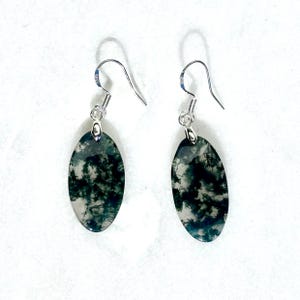 May include: A pair of oval-shaped moss agate earrings featuring dark green, tree-like inclusions within a translucent stone. Each pendant is suspended from a silver-toned metal hook with a small loop attachment, showcasing a polished finish.