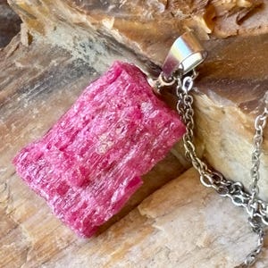 May include: A raw, deep pink crystal pendant with a rough, textured surface and vertical striations. The stone is secured by a silver-toned metal bail and hangs from a matching silver-toned metal chain, showcasing a natural, unpolished aesthetic.