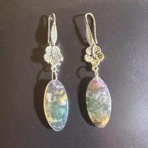 May include: A pair of dangle earrings. Each earring features a silver-toned floral accent and a long, oval-shaped stone with green, yellow, and purple hues. The earrings have a hook closure.