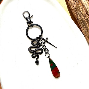Peut inclure: Un porte-clés en métal noir avec un fermoir mousqueton et un anneau circulaire. Trois breloques sont fixées à l'anneau : un serpent noir texturé, une petite épée noire et une pierre lisse en forme de goutte aux motifs verts et rouges.