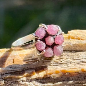 Puede incluir: Un anillo de plata con un grupo de gemas rosas. Las gemas están facetadas y dispuestas en un patrón floral. El anillo está sobre un fondo de madera natural, con la luz del sol resaltando los detalles.