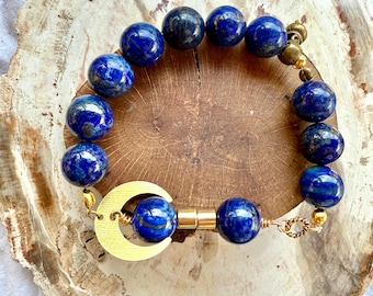 May include: A bracelet with deep blue lapis lazuli beads, featuring a gold-coloured crescent moon charm and gold-toned metal details. The bracelet is displayed on a wooden surface.