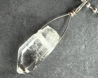 Könnte beinhalten: Ein klarer Quarz-Kristall-Anhänger mit silbernem Draht und einer Kugelkette. Der Kristall hat eine helle, durchscheinende Farbe und eine spitze, geometrische Form. Die Halskette wird auf einer dunkelgrauen Oberfläche präsentiert.