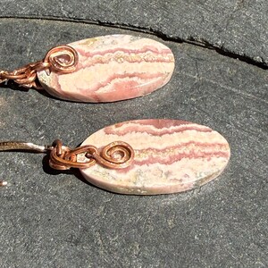 May include: A pair of oval earrings. Each earring features a pink and white stone with a striped pattern, accented with copper wire detailing. The copper wire is shaped into a spiral at the top of each earring.