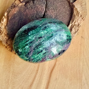 Pode incluir: Uma pedra preciosa oval polida, predominantemente verde com veios pretos e castanhos. Descansa sobre uma superfície de madeira, ao lado de um pedaço de madeira castanha escura texturizada. A forma da pedra é lisa e arredondada.