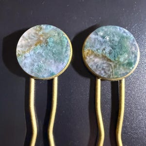 May include: Two hair pins with round, moss agate stones in brass settings. The stones feature a mix of green, white, and gold colors. The brass pins have a curved design.