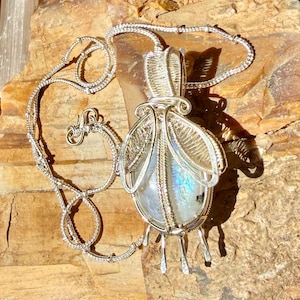 May include: A silver wire-wrapped pendant featuring an oval moonstone with blue iridescence, suspended on a matching silver chain. The metalwork includes intricate coiled wire details forming wing-like structures and a secure bail at the top.