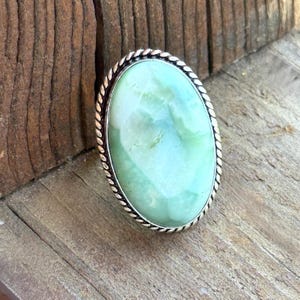 May include: An oval-shaped ring featuring a smooth, light green stone set in a polished silver-toned metal frame. The setting is characterized by a distinct twisted rope-like texture that encircles the perimeter of the large, pale green cabochon.