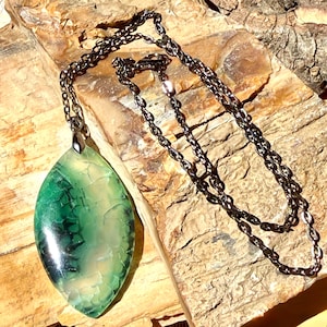 May include: A marquise-shaped pendant featuring a translucent green stone with a cracked, webbed texture, suspended from a dark metallic chain. The stone displays varying shades of emerald and light green with dark, vein-like inclusions throughout.