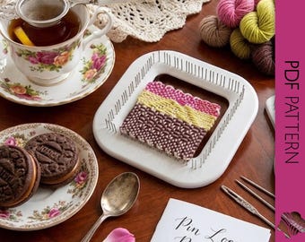 May include: A cozy scene featuring a teacup with a floral design, a plate of cookies, and a pin loom with a colorful woven sample. Yarn skeins and a booklet titled "Pin Loom Patterns" are also present, suggesting a crafting theme.