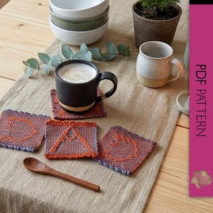 May include: A table setting with a black mug of coffee on a knitted coaster. Three additional coasters with orange and blue geometric designs are visible. A wooden spoon and a white mug are also present.