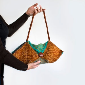 May include: A woven handbag in shades of brown, teal, and orange. The bag features a unique geometric shape with a flap secured by a large, round wooden button. Braided brown straps extend from the top of the bag.