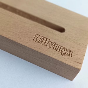 May include: A light-colored wooden phone stand with a carved slot and the word "Ulvunga" etched into the wood. The stand is rectangular and sits on a white surface.