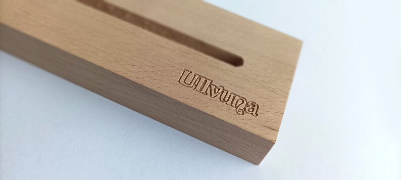 May include: A light-colored wooden phone stand with a carved slot and the word "Ulvunga" etched into the wood. The stand is rectangular and sits on a white surface.