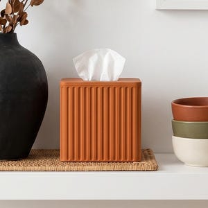 May include: A decorative shelf arrangement featuring a black vase with dried branches, a terracotta tissue box, and a stack of three ceramic bowls in earth tones. A gold candle holder and framed art complete the display.